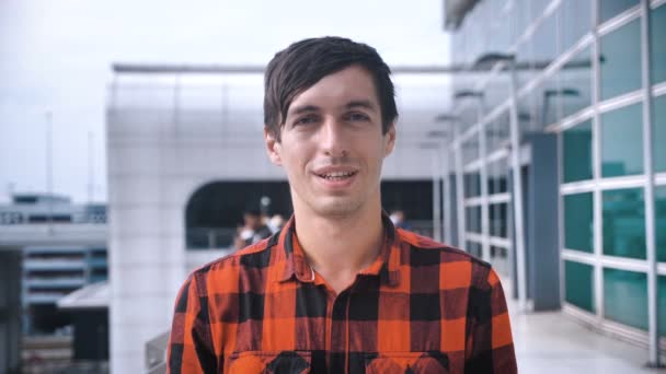 Portret van een jonge man in een geruit hemd glimlachend kijkend naar de camera buiten op een grote moderne stedelijke achtergrond - Video