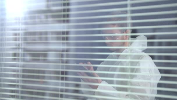 Άνδρας με λευκά προστατευτικά ενδύματα εργασίας και γυαλιά σταθεί με τηλέφωνο στο εργαστήριο - Πλάνα, βίντεο