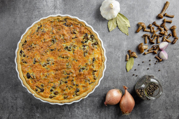 Traditional french pie Quiche Lorraine with mushrooms, potatoes, cheese, onions and seasonings. In a white baking dish, with forks, with black pepper and bay leaf, on a concrete background.Top view. - Photo, Image