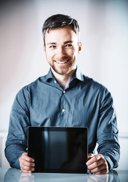 Young man with a beaming friendly smile holding up a blank tablet-pc with the screen to the viewer for your advertising or text - Foto, imagen