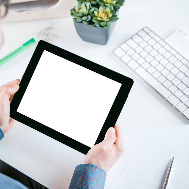 Podnikatel drží tablet s bílou prázdnou obrazovkou v rukou s kopírovacím prostorem pro vaši reklamu nebo text, vysoký úhel pohledu na jeho ruce přes jeho stůl - Fotografie, Obrázek