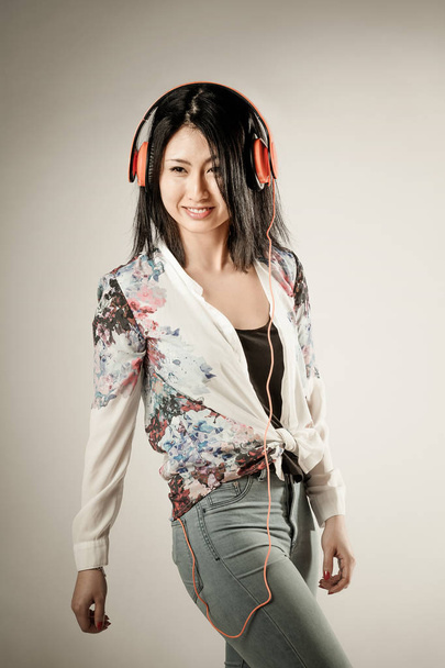 Vivacious trendy slender young Asian woman with a charismatic playful smile enjoying her music on a set of stereo earphones over a studio background with a vignette - 写真・画像
