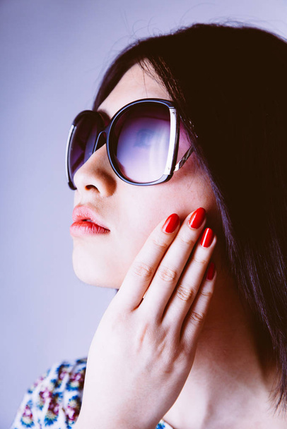 Fashionable gorgeous young Asian woman in trendy sunglasses with her hand to her cheek showing off her manicured nails looking to the side with a serene expression - Fotó, kép
