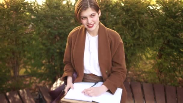 Short-haired girl with notebooks and abstracts in a brown coat. A student is preparing for exams while sitting on a bench in a park - 映像、動画