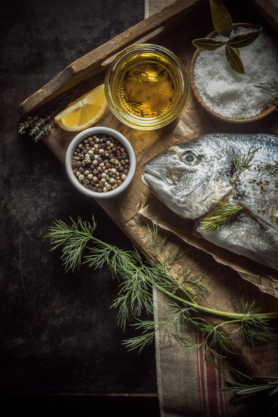 Uncooked whole fresh fish with herbs, olive oil and spices for marinating and rubbing lying on a vintage wooden tray in a rustic country kitchen, overhead view with copyspace - 写真・画像
