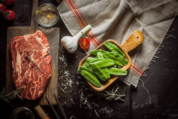Still Life of Raw Ingredients for Preparing Dinner Meal - Raw Steak on Cutting Board, Snow Peas in Wooden Dish, Garlic Bulb and Various Herbs and Spices and Hand Towel - Фото, зображення