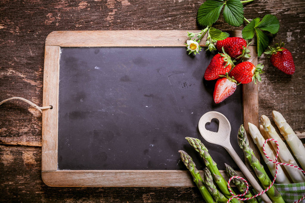 Small Empty Black Chalkboard with Fresh Asparagus and Strawberries on the Edge, Placed on Rustic Table, Emphasizing Copy Space. - Foto, Bild