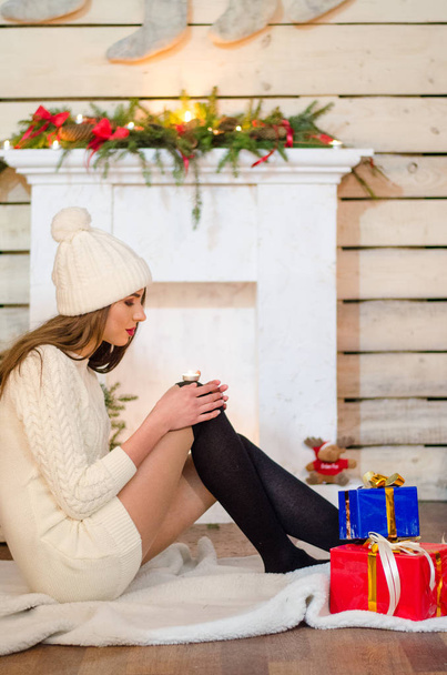 Hermosa mujer sexy con árbol de Navidad en el fondo sentado en una silla elegante en un paisaje acogedor. Retrato de niña posando vestido blanco ajustado bastante corto. Atractiva morena femenina, tiro de interior
. - Foto, imagen