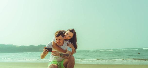 Happy couple taking selfies near sea. Loving couple embracing during date on beach against waving sea and cloudless sky. - Fotoğraf, Görsel