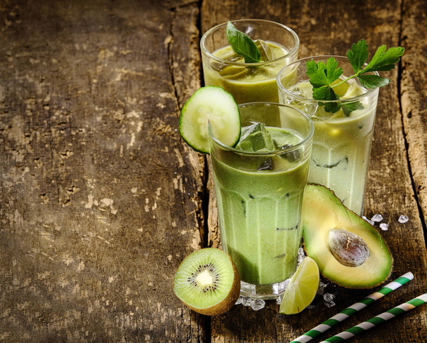Three Tall Glasses Filled with Healthy Green Smoothie Shake Make from Avocado, Kiwi, Cucumber and Lime, Garnished with Herbs and Ice, on Wooden Surface with Raw Ingredients and Copy Space - 写真・画像