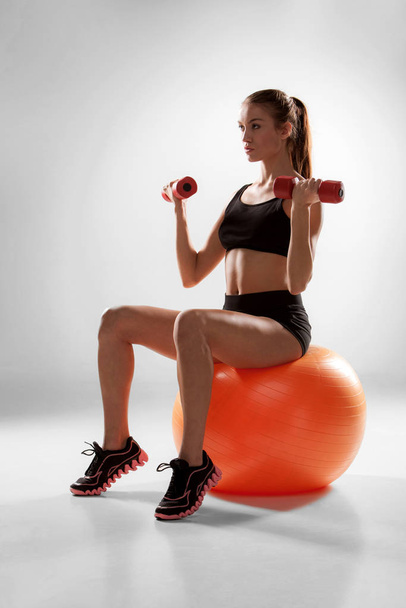 Sporty woman doing aerobic exercise with red dumbbells on a fitness ball on grey background - Photo, Image