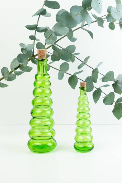 Glasgrüne Flaschen und ein Eukalyptuszweig auf weißem Hintergrund. Ätherisches Eukalyptusöl für den Wellnessbereich. - Foto, Bild