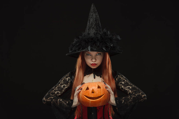 Mädchen im Hexenkostüm mit Halloween-Kürbis isoliert auf schwarzem Hintergrund - Foto, Bild