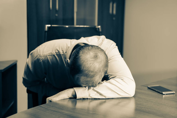 Muž středního věku sedí za stolem v kanceláři, sklíčený a vyčerpaný. Jen sedí na pracovišti s hlavou na stole. Sepia barva a vodorovný obrázek. - Fotografie, Obrázek