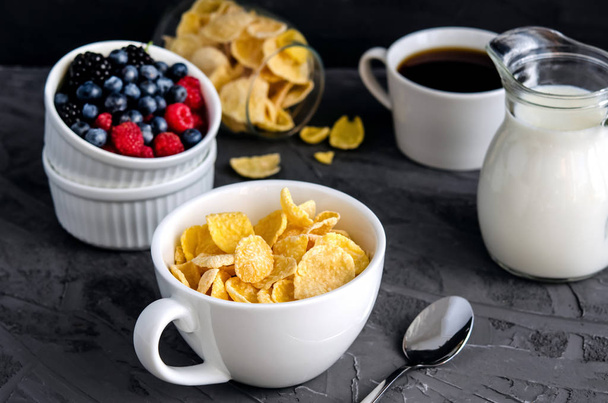 Healthy breakfast with cornflakes in a white cup, berries, milk and coffee on a dark gray background. - Photo, image