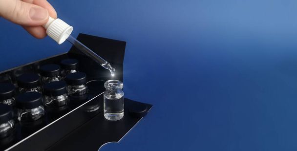 Fingers holding pipette with sparkling drop flowing out, measuring cosmetic product in glass flacon, set of vials in black box behind, on blue background.Cosmetic brand packaging, dosing.Copy space
 - Фото, изображение