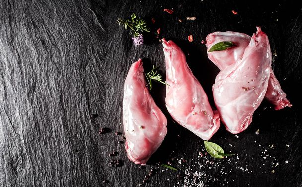 High Angle View of Raw Rabbit Meat, Rear Leg Cuts of Hare Seasoned with Fresh Herbs and Spices on Dark Textured Surface with Copy Space - Zdjęcie, obraz