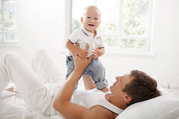 Young smiling father with his nine months old son on the bed at home on white home background - Φωτογραφία, εικόνα