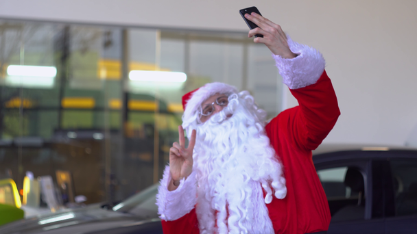 Santa Claus tomando una selfie en un concesionario de automóviles y mostrando el gesto "paz
" - Imágenes, Vídeo