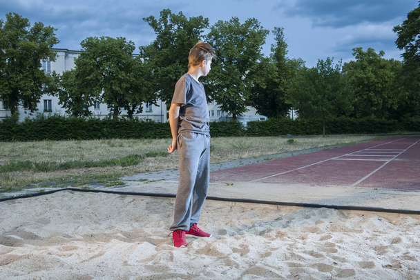 full length side view of a young male teenager standing in a jump pit standing on the run-in stretch with arms folded on his back - Fotografie, Obrázek