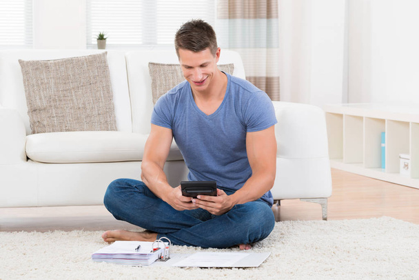 Young Man Sitting On Carpet Calculating Budget With Calculator - Foto, immagini