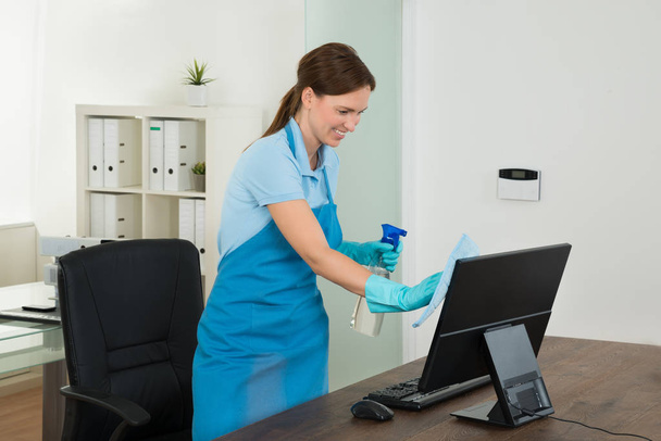 Young Happy Female Janitor Cleaning Wooden Desk With Rag In Office - Foto, Bild