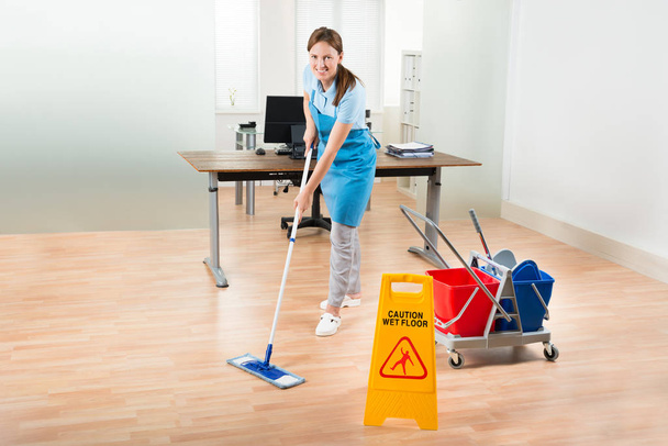 Happy Female Janitor With Cleaning Equipments Cleaning Hardwood Floor In Office - 写真・画像