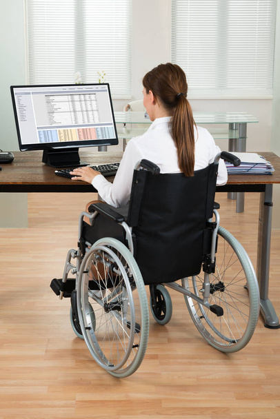 Young Female Architect On Wheelchair Looking At Blueprint On Computer In Office - Foto, imagen