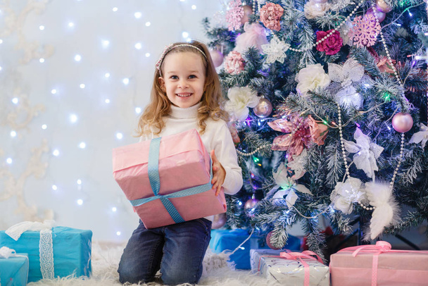 Happy pretty girl with gift boxes sits near Christmas tree in the room of the house.  Merry Christmas and New Year. - Photo, Image