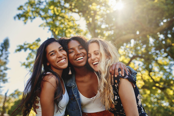 Three smiling multiethnic young women in casual wear standing together under the bright sunlight at the park having fun and laughing - Фото, изображение