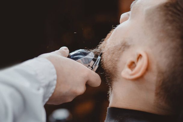 Крупный план парикмахера стригущей бороду мужчине в парикмахерской электрической бритвы - Фото, изображение