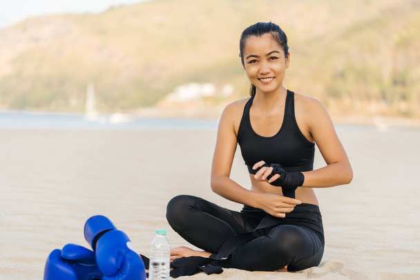Azjatka w sportowej odzieży siedząca na plaży i trzymająca się za ręce przed treningiem bokserskim. Zdrowy styl życia i letni nastrój. Portret z bliska z uśmiechem. - Zdjęcie, obraz