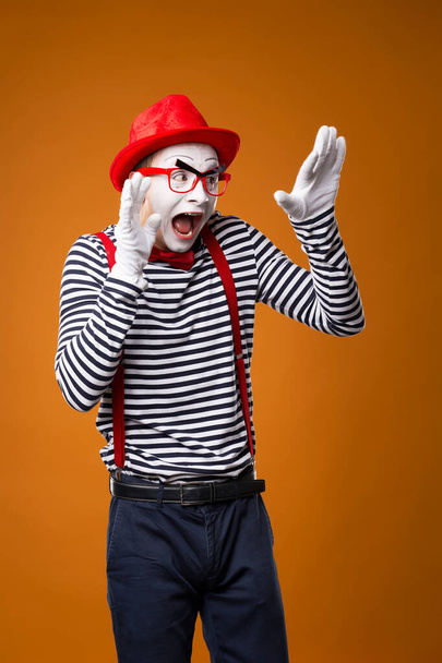 Frightened mime man in vest and red hat on orange background. - Фото, изображение