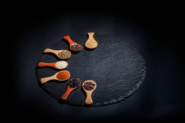 Different spices, peppers, coriander, sesame seeds and others for cooking are in wooden spoons on a black background - Zdjęcie, obraz