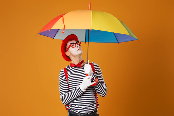 Mime looking up with multi-colored umbrella in hand isolated on blank orange background - Fotografie, Obrázek