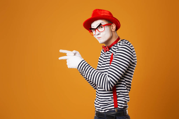 Mime in rode hoed en in vest wijzend met zijn hand naar zij op oranje achtergrond - Foto, afbeelding
