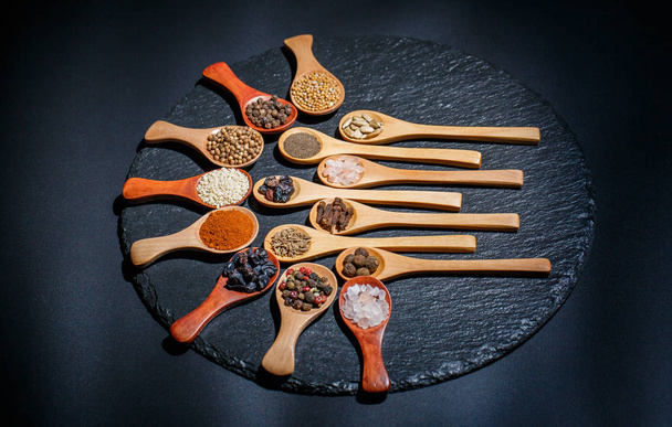 Different spices, peppers, coriander, sesame seeds and others for cooking are in wooden spoons on a black background - Zdjęcie, obraz