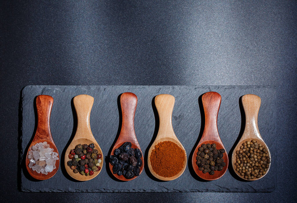 Diferentes especias, pimientos, cilantro, semillas de sésamo y otros para cocinar están en cucharas de madera sobre un fondo negro
 - Foto, imagen