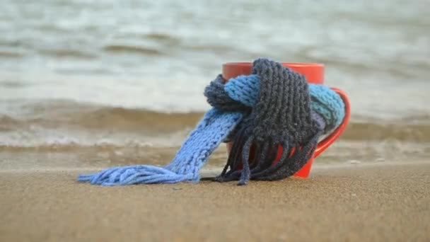 Red mug with coffee tied with blue knitted scarf stands on sandy beach - Footage, Video