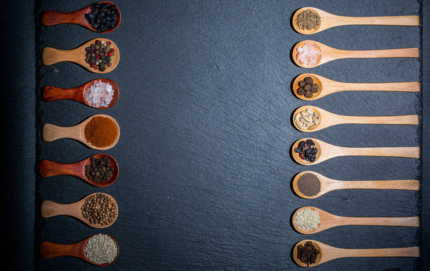 Different spices, peppers, coriander, sesame seeds and others for cooking are in wooden spoons on a black background - Foto, Imagem