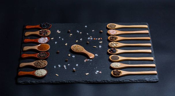 Different spices, peppers, coriander, sesame seeds and others for cooking are in wooden spoons on a black background - Foto, imagen
