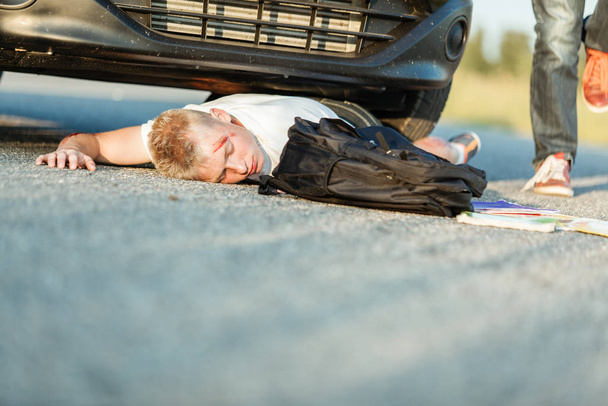 Young schoolboy knocked over by a car in a traffic accident lying face down on the tarmac with bleeding injuries and his school bag alongside him - Фото, изображение