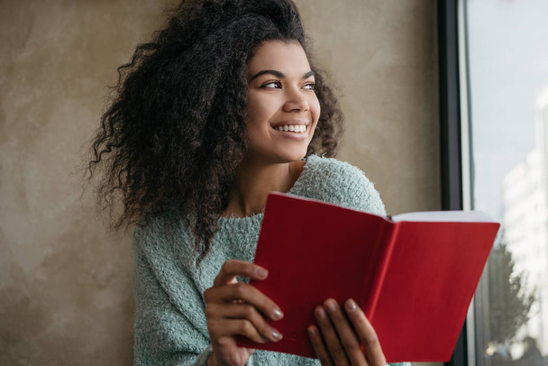 Successful university student studying, learning language, sitting at library. Portrait of beautiful African American woman holding red book, looking at window and smiling. Exam preparation concept - Foto, Imagen