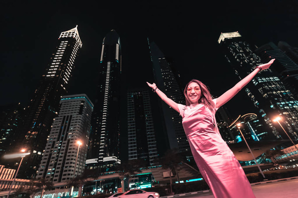 Feliz mujer asiática en vestido levantando las manos delante de los rascacielos en la ciudad de la noche
 - Foto, imagen