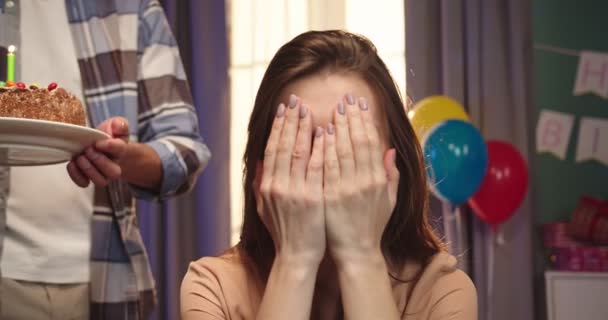 Portrait of the beautiful caucasian young woman sitting and closing eyes with hands while her handsome husband bringing birthday cake and she blowing candles. - Filmmaterial, Video