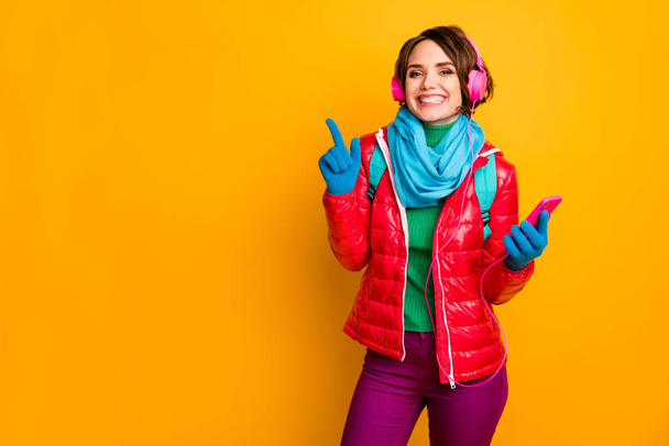 Photo of funny blogger lady hold telephone listen earphones directing finger empty space wear casual red coat blue scarf gloves trousers isolated yellow color background - Foto, afbeelding