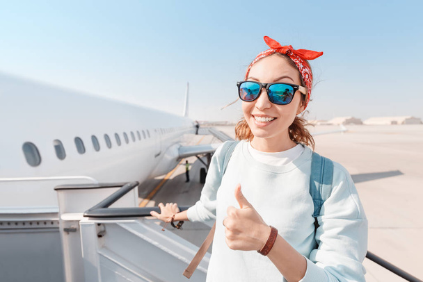 Ένα νεαρό κορίτσι χαμογελάει και σηκώνει τους αντίχειρες ενώ επιβιβάζεται σε ένα μεγάλο λευκό αεροπλάνο στο αεροδρόμιο. Έννοια του ταξιδιού και νίκη κατά της αεροφοβίας - Φωτογραφία, εικόνα
