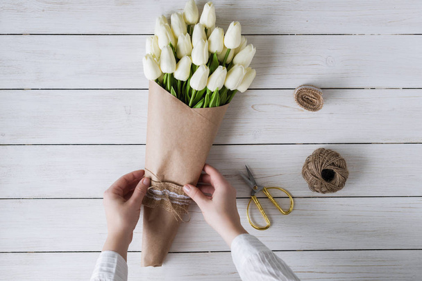 Woman florist wrapping beautiful bouquet of white tulips in pack craft paper on the wooden table. Flat lay - Фото, изображение