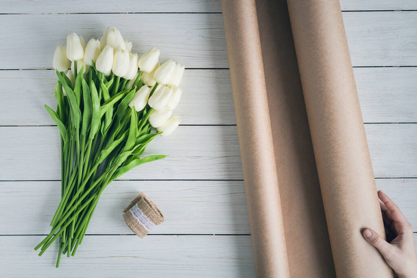 Woman florist wrapping beautiful bouquet of white tulips in pack craft paper on the wooden table. Flat lay - 写真・画像