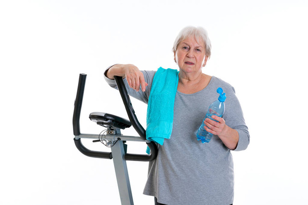 gray-haired female senior with water bottle train with fitness machine - Zdjęcie, obraz